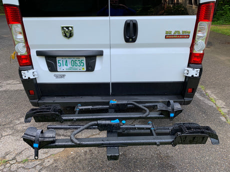 RV rental: Rear of a white Ram ProMaster van with a two-tier bicycle hitch rack, ready for adventure on a wet day.