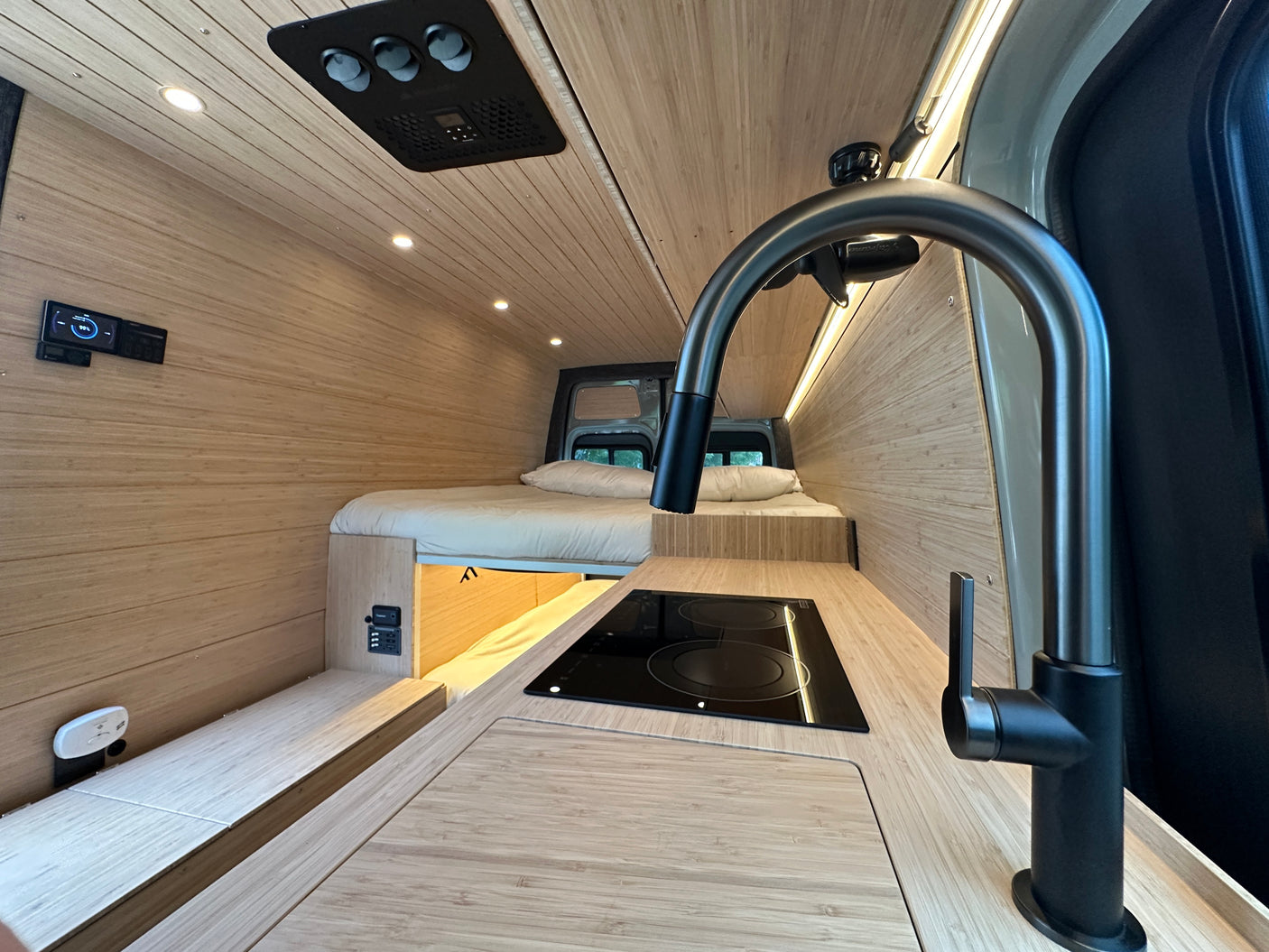 Modern rv rental interior: light wood paneling, kitchen with faucet and cooktop, comfy bed, and control panels for a cozy trip.
