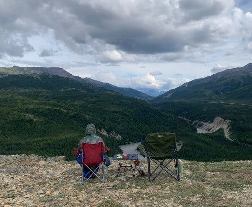 A person in a red chair overlooks a vast mountain valley with a river, perfect for an RV rental escape.