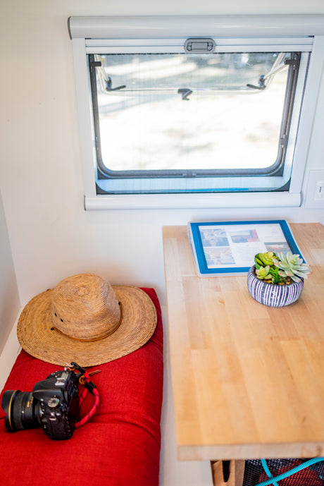 Inside an RV rental: a window, a red bench with a straw hat and camera, plus a wooden table with a binder and plant.