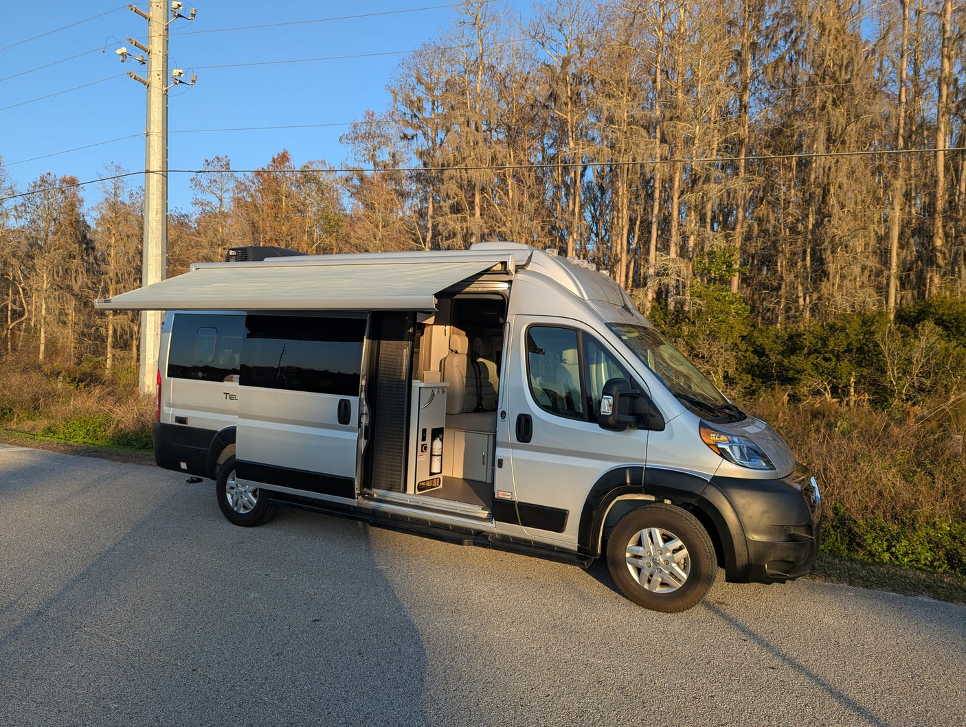 Silver camper van RV rental parked on a sunny road. Awning extended, side door open showing interior, ideal for outdoors travel.