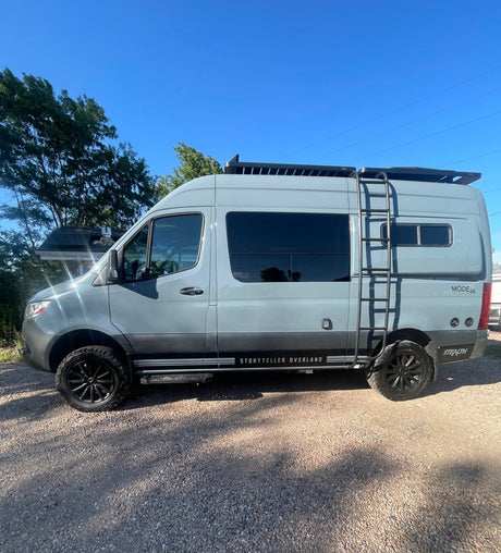 This gray Storyteller Overland Mode 4x4 camper van, ideal for rv rental, is parked on gravel with blue sky & trees.