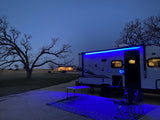 Enjoying the evening at an RV rental campsite. Blue lights glow on the trailer, outdoor rug, table & chairs.