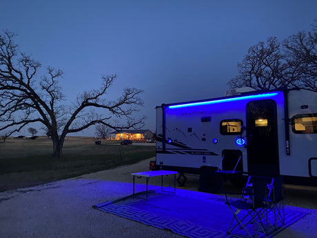 Enjoying the evening at an RV rental campsite. Blue lights glow on the trailer, outdoor rug, table & chairs.
