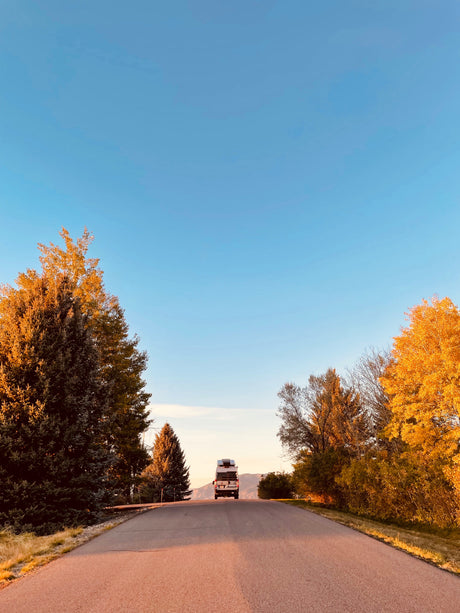 A white RV journeys up a reddish road, flanked by golden autumn trees and distant mountains under a clear blue sky. Ideal for an RV rental adventure.