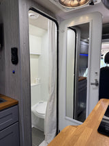 RV rental interior: compact bathroom with toilet & shower curtain. A mirror on the pivoting door reflects wooden counters & the main cabin.