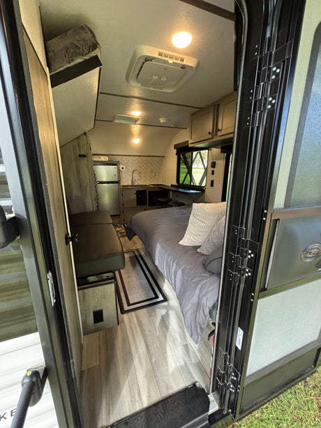 View into a compact RV rental camper, showing a cozy bed, kitchenette, dinette, and ceiling AC unit.