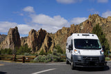 Scenic view: a white camper van, perfect for RV rental, parked amidst towering rock formations under a blue sky.