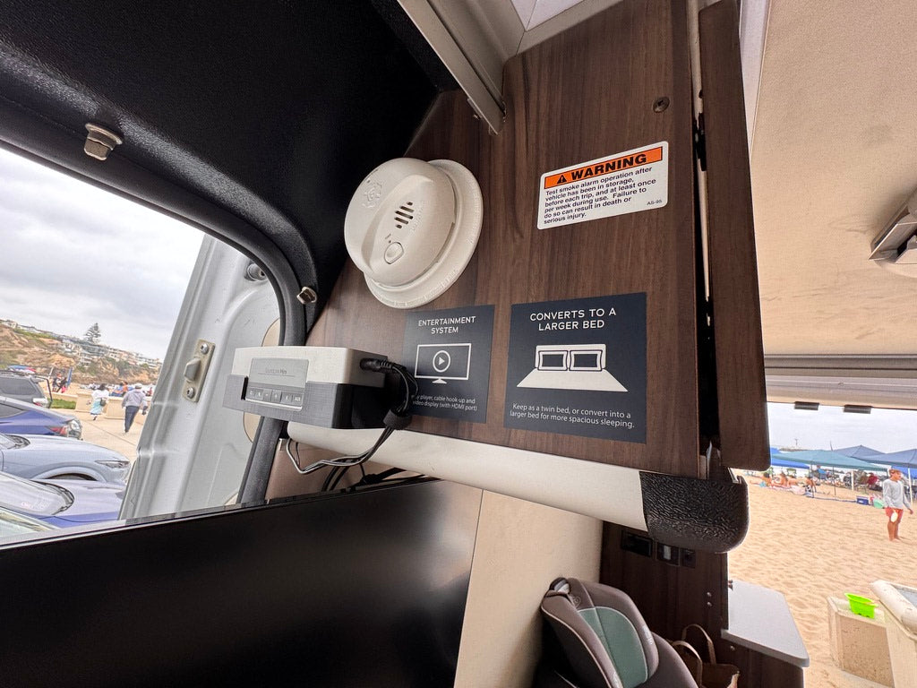 RV rental interior: smoke alarm, retro entertainment console, and bed conversion guide. Outside, a lively beach view.