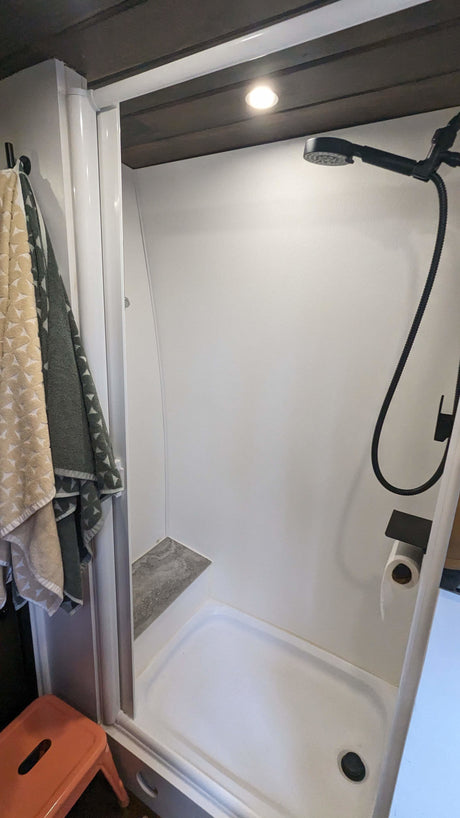 Inside an RV rental: a clean, compact shower with a handheld sprayer, corner shelf, and essential amenities.