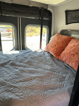 A comfortable bed with a gray quilted cover and orange pillows inside an rv rental, with sunny outdoor views.