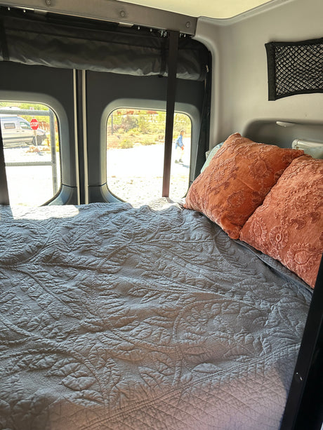 A comfortable bed with a gray quilted cover and orange pillows inside an rv rental, with sunny outdoor views.