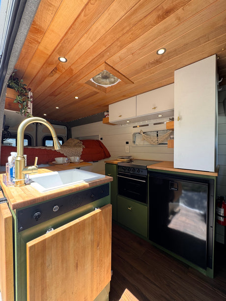 Stylish camper van kitchen interior for your next rv rental, featuring a sink, oven, fridge, and cozy bed in background.