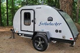 A small, white Bushwhacker teardrop camper, ready for an RV rental adventure. This compact travel trailer is parked on dirt in a forest.