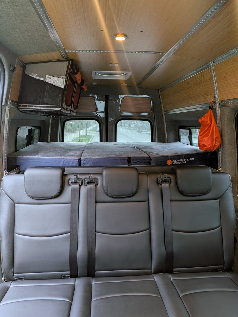 This rv rental interior features a 3-person black leather seat, custom bed with "Adventure Wagon" logo, wood-paneled ceiling, & windows showing a rainy day. — photo 3