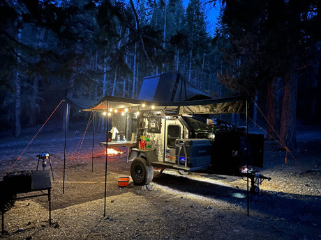 An off-road trailer RV rental lights up a cozy nighttime campsite in the forest, featuring a warm campfire and outdoor gear.