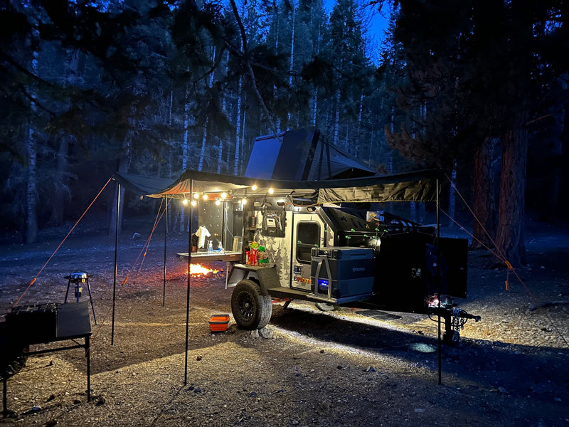 An off-road trailer RV rental lights up a cozy nighttime campsite in the forest, featuring a warm campfire and outdoor gear. — photo 18
