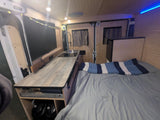 Interior of an RV rental showcasing a bed, compact kitchen with sink/cooktop, and TV. Features wood paneling and bright lights.
