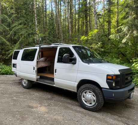 White Ford camper van RV rental in a forest, side door open to a cozy interior bed. Ready for adventure.
