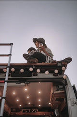 Rooftop relaxation on a camper van RV rental. A woman in a hat sips wine amidst glowing string lights, embracing van life travel.
