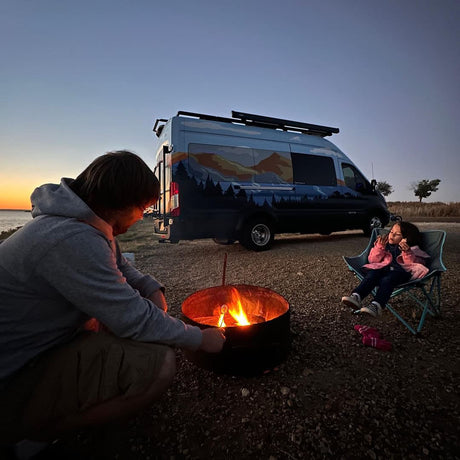 Family camping at dusk by an rv rental van. A man tends the campfire while a child watches. Outdoor adventure.