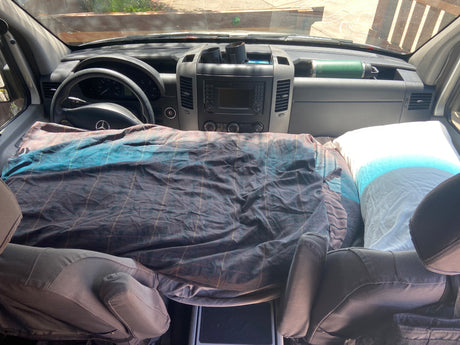 RV rental interior: a cozy bed with plaid bedding fills the front cabin, featuring a Mercedes dashboard and Yeti bottle.