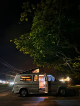 Night scene: a spacious pop-top RV rental with interior lights on and door open, parked under a large tree.