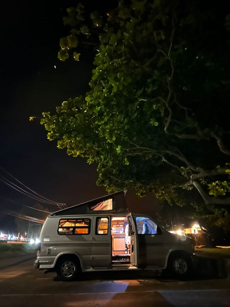 Night scene: a spacious pop-top RV rental with interior lights on and door open, parked under a large tree.