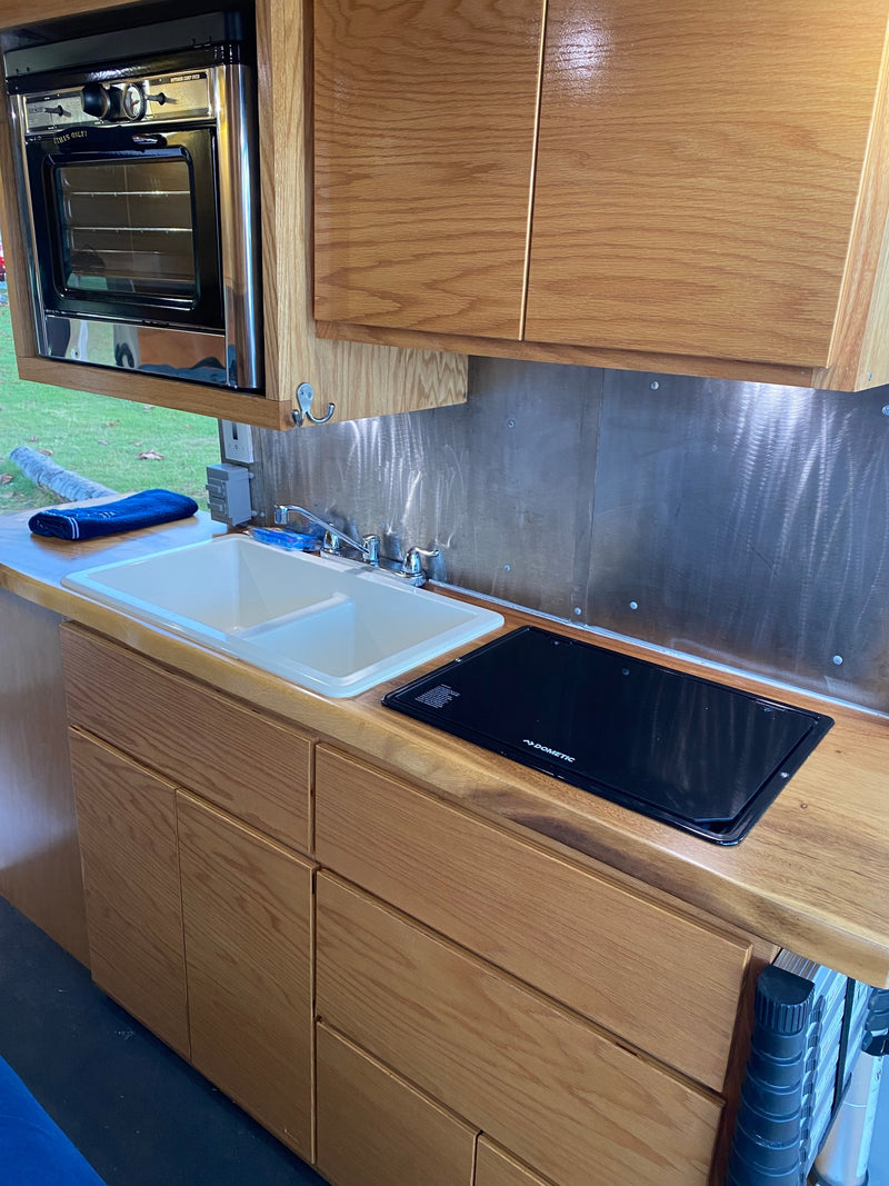 Compact RV rental kitchen with a dual sink, Dometic cooktop, and a built-in outdoor camp oven, complemented by wood cabinets. — photo 18