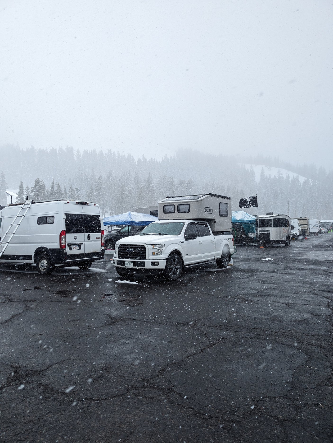 Snow-covered lot with diverse RVs like a white camper van, truck w/ Scout camper, and trailers. Ideal for winter rv rental adventures.