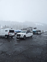 Snow-covered lot with diverse RVs like a white camper van, truck w/ Scout camper, and trailers. Ideal for winter rv rental adventures.