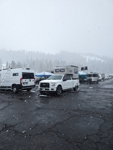 Snow-covered lot with diverse RVs like a white camper van, truck w/ Scout camper, and trailers. Ideal for winter rv rental adventures.