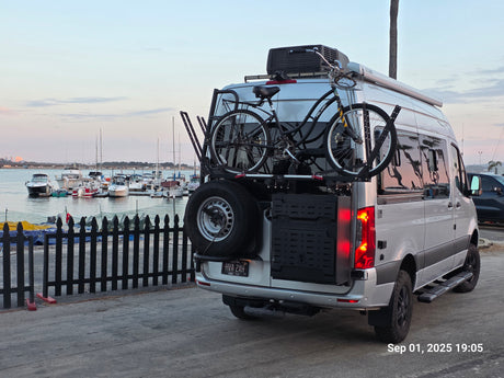 White Sprinter RV rental with bikes & spare tire at a marina. Ideal for travel & outdoor adventure.