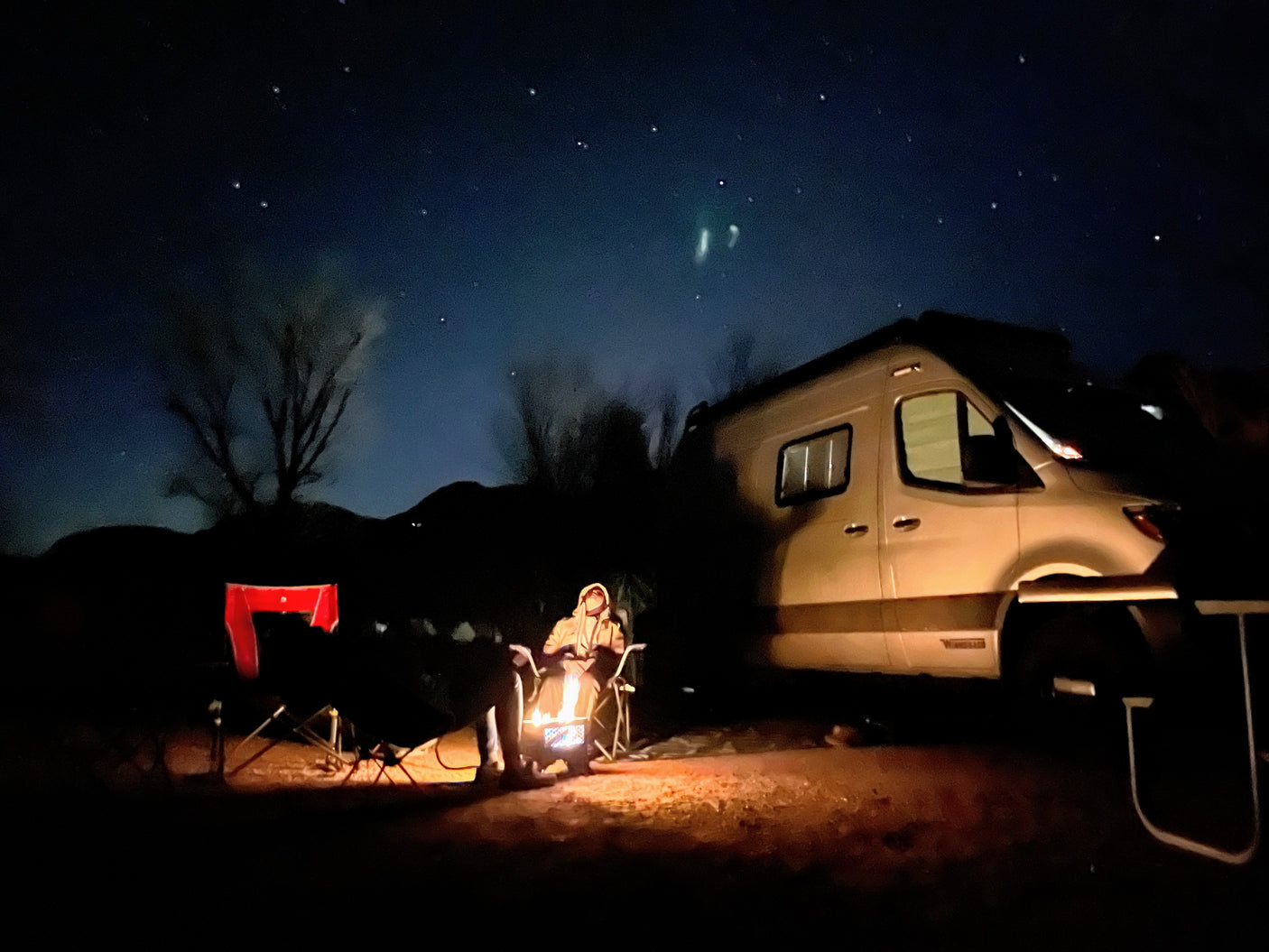 Starry night camping in an RV rental! A person sits by a glowing fire, near a modern camper van under a clear sky.