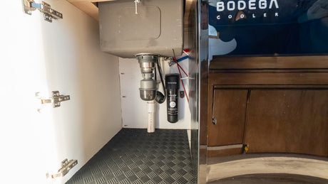 An RV rental's under-sink area, featuring plumbing, a Waterdrop filter, and a durable floor mat for a compact kitchen.