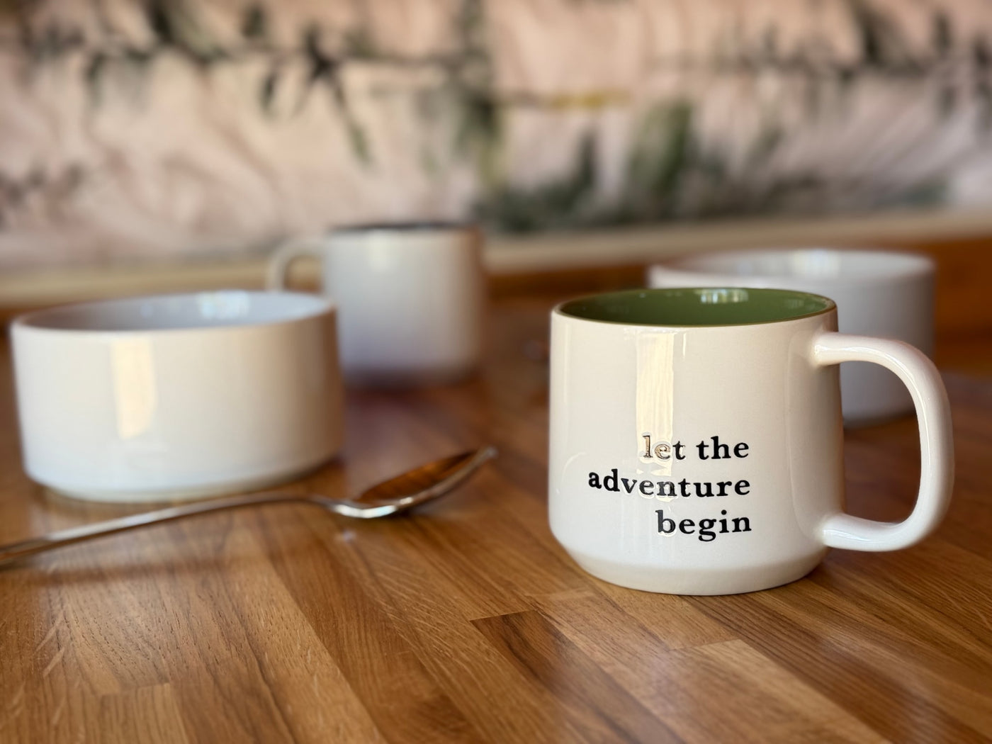 A white mug with "let the adventure begin" sits on a wooden table, with other cups and a spoon nearby. Perfect for your RV rental trip! — photo 5