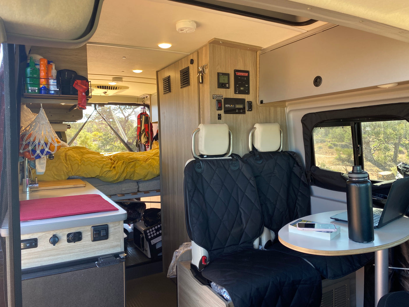 Interior of an RV rental: a compact living space with kitchen, bed, and a driver's area with control panels.