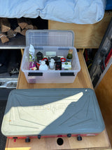 RV rental kitchen pull-out featuring Camp Chef stove, food bin with essentials, and stored firewood. Ready for van life!
