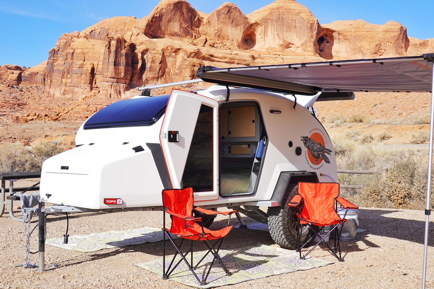 White teardrop camper RV rental with awning & open door, set in a scenic red rock desert. Camping chairs wait by the entrance. — photo 2