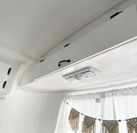 White RV rental interior showing overhead storage, a bright ceiling light, and cozy sheer curtains with macrame decor.