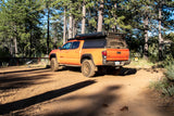 An orange Toyota Tacoma overland vehicle, functioning as an RV rental, parked amidst tall pines for camping.