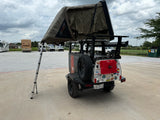 An equipped camping trailer with a 23ZERO rooftop tent displayed at an RV rental lot, surrounded by RVs and rental vans.