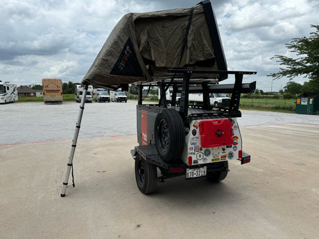 An equipped camping trailer with a 23ZERO rooftop tent displayed at an RV rental lot, surrounded by RVs and rental vans.