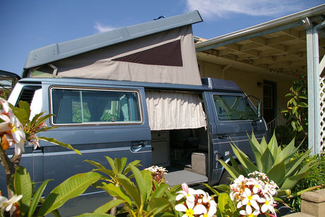 Blue pop-top camper van with open doors, ready for an RV rental adventure. Lush flowers frame this cozy home on wheels.