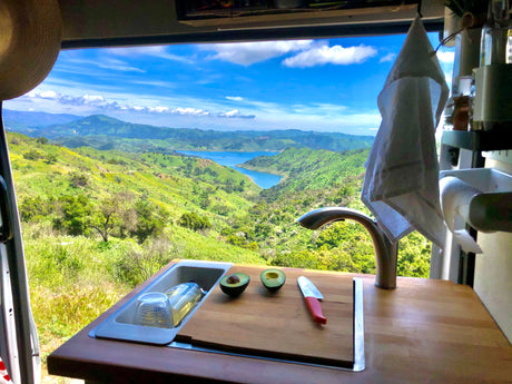 Inside an RV rental: a kitchen counter with avocado & sink, showcasing a panoramic view of lush green mountains and a blue lake.