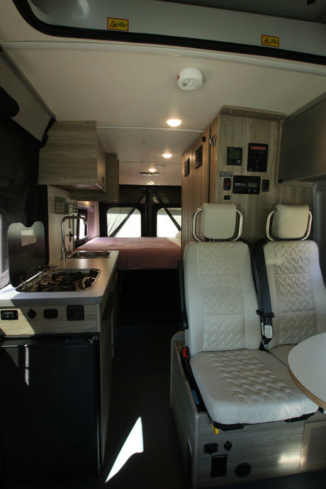 Interior of an RV rental showing a full kitchenette on the left, a bed at the back, and front passenger seats on the right.