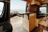 Open side door of an RV rental revealing a cozy wooden interior with kitchen, facing a sandy beach and ocean waves.