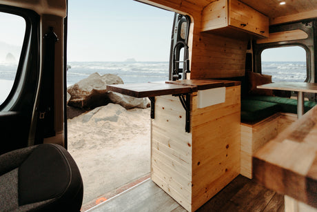 Open side door of an RV rental revealing a cozy wooden interior with kitchen, facing a sandy beach and ocean waves.