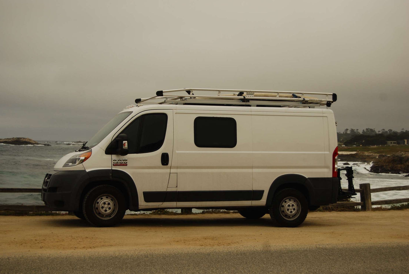 White Ram ProMaster RV rental van with roof rack parked at a scenic coastal spot by crashing ocean waves.