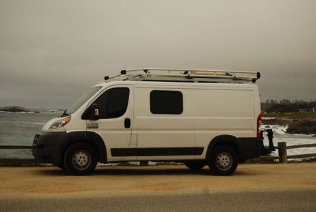 White Ram ProMaster RV rental van with roof rack parked at a scenic coastal spot by crashing ocean waves.